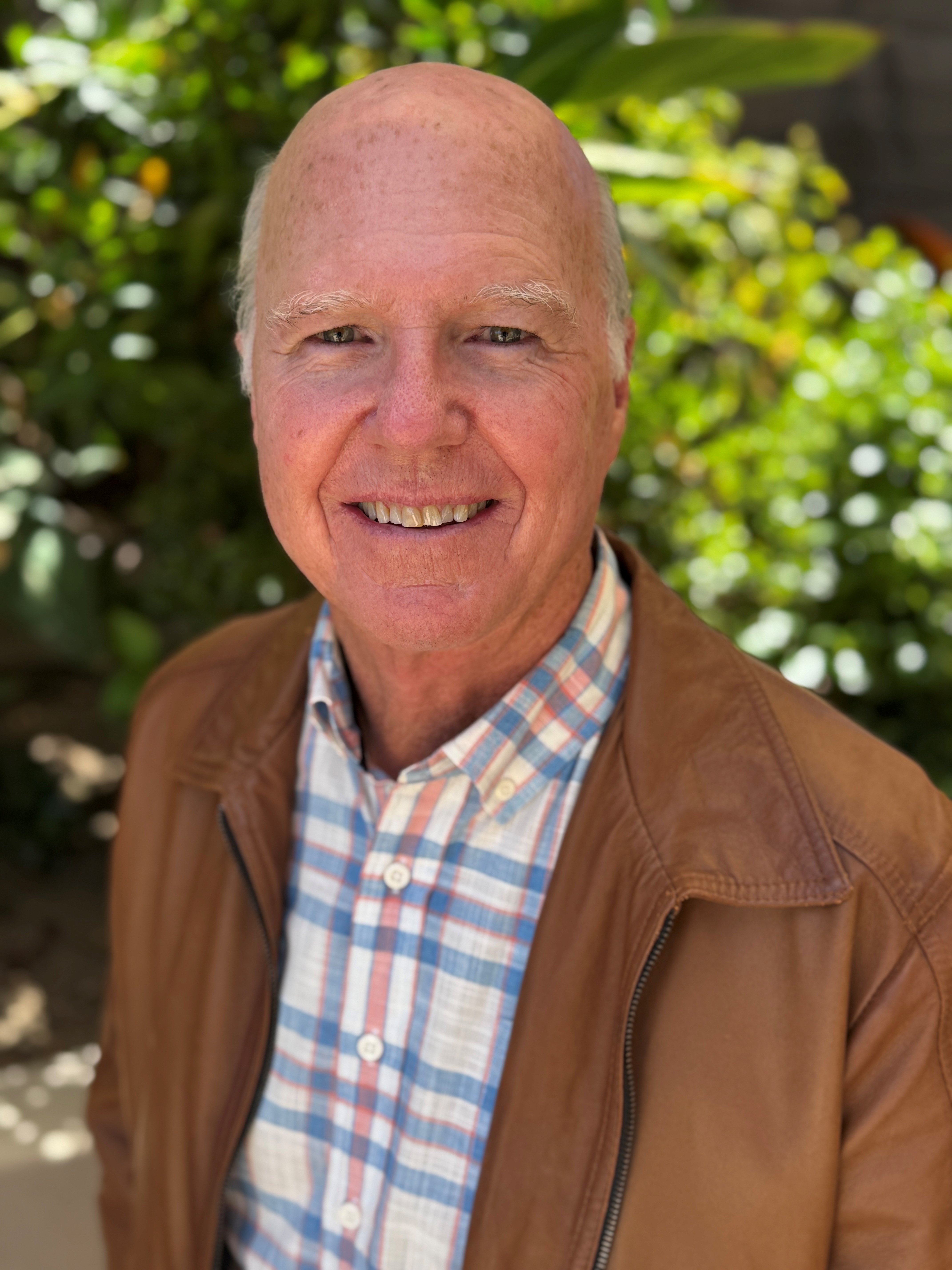 Don Mitchell smiling outdoors, wearing a brown jacket over a plaid shirt, stands in front of lush green foliage. The mood is warm and cheerful.