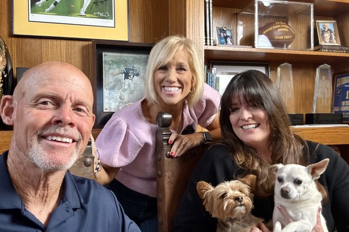 John and Holly Carney smiling together in front of shelves of football memorabilia and trophies.