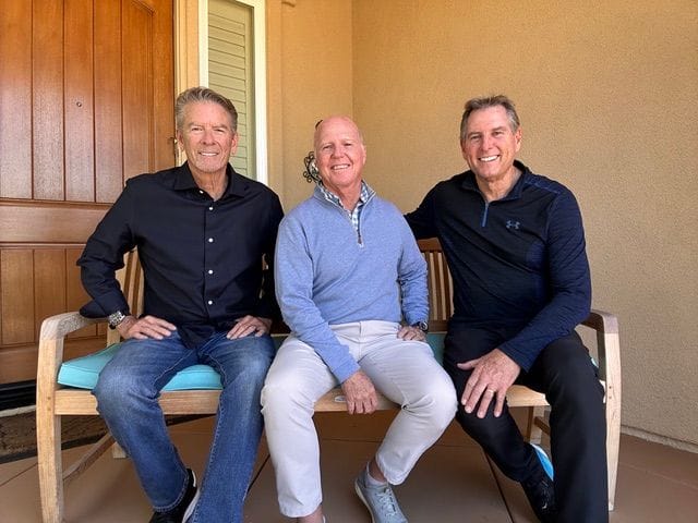 Three men sitting side by side on a wooden bench on a sunlit porch, smiling warmly at the camera in relaxed conversation.